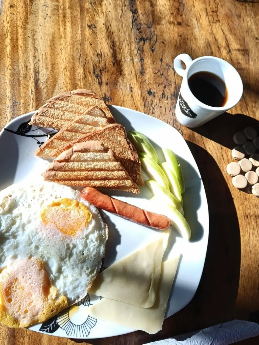 Desayuno sobre una mesa de madera: huevos fritos, pan tostado, salchicha, queso y una taza de café negro.