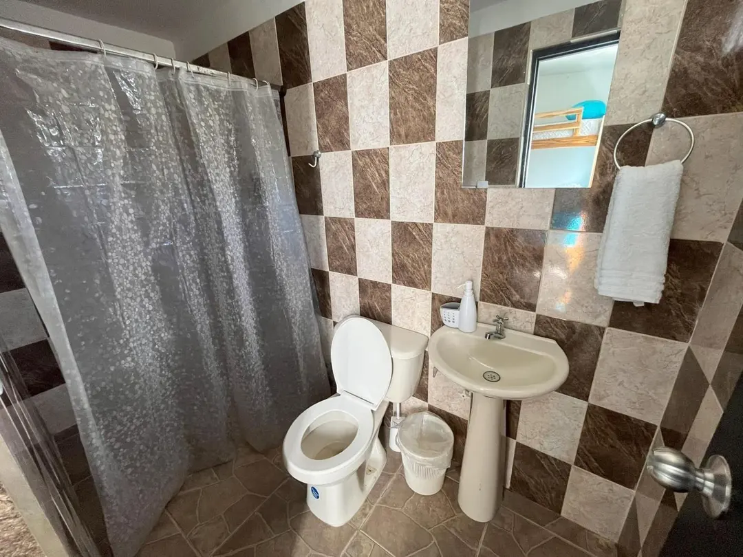 Baño privado con ducha de cortina, sanitario, lavamanos de pedestal y paredes con baldosas en damero café y blanco.
