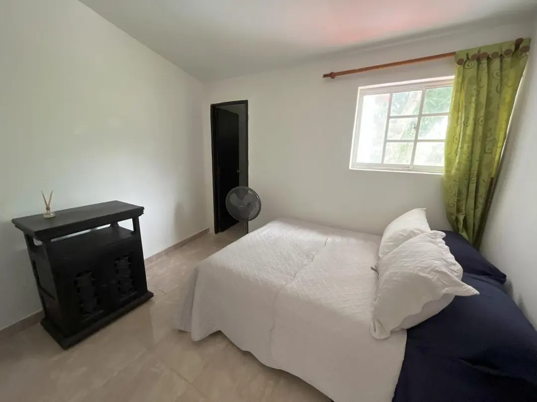 Cama doble con cobija blanca, una cómoda oscura y una ventana con cortina verde.