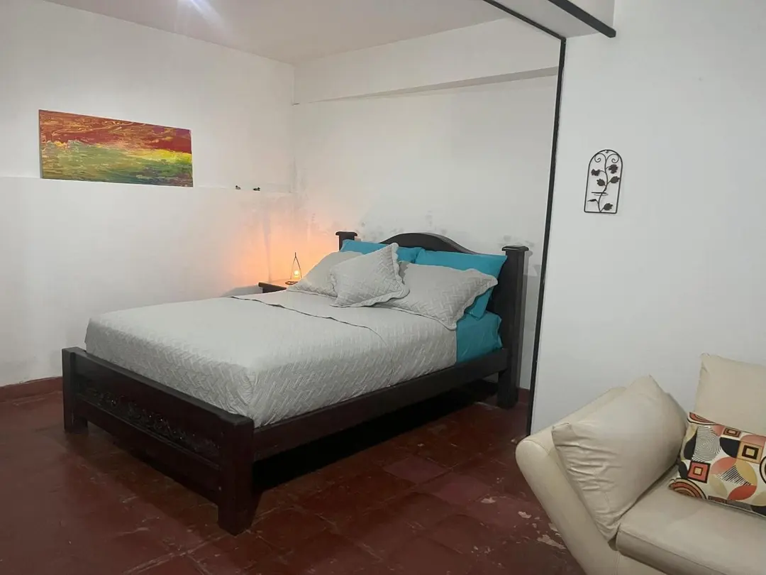 Cama doble con sábanas turquesa, un cuadro de tonos cálidos en la pared y un sofá claro.