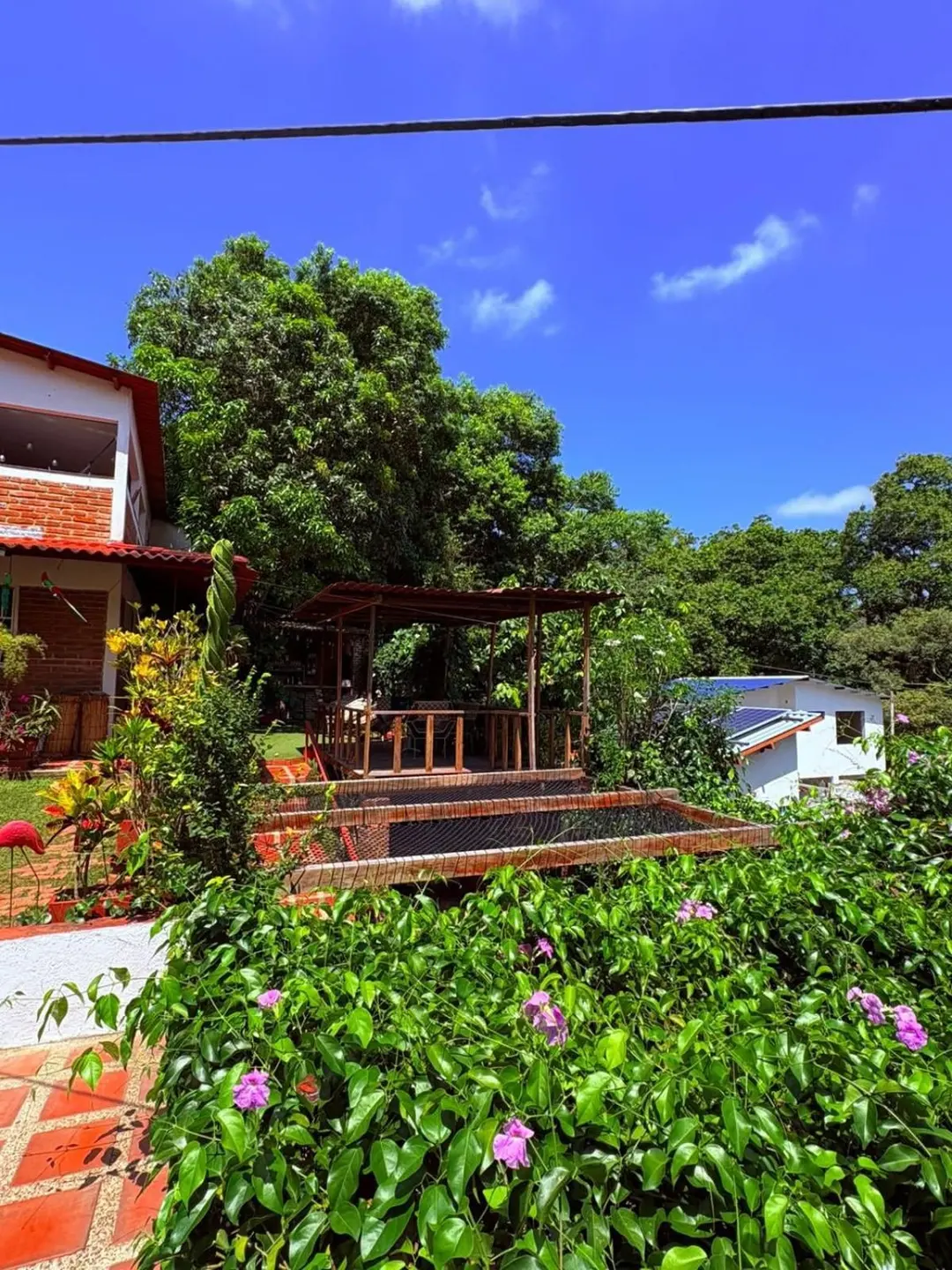 Arbustos con flores rosadas en primer plano; detrás, el kiosco y la casa bajo un cielo azul.