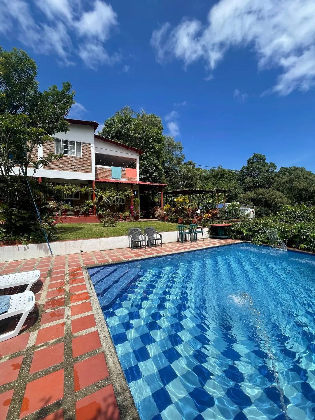La piscina y la casa bajo un cielo azul con nubes, con el borde de ladrillo en primer plano.