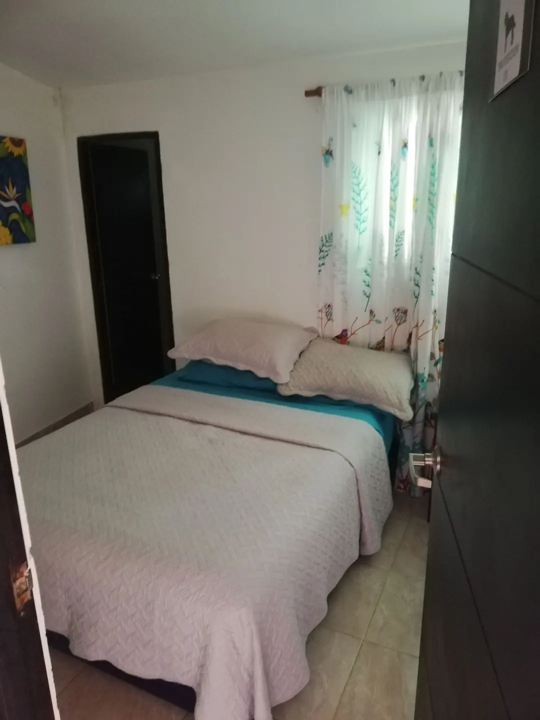 Habitación privada con cama doble de sábanas turquesa, cortinas blancas con hojas estampadas y puerta oscura.