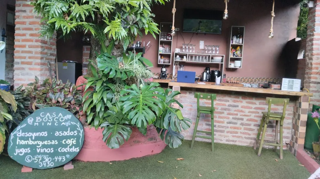 La barra de la casa con plantas y el tablero de tiza verde: Casa Bosco Minca, desayunos, asados, hamburguesas, café, jugos, vinos, cocteles y un número de WhatsApp.
