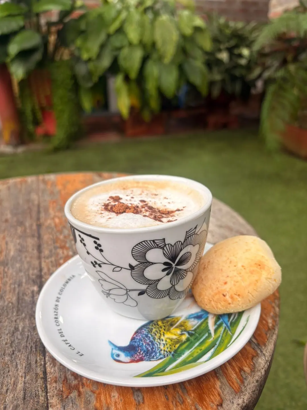 Capuchino con cacao espolvoreado en una taza de flores y un pan redondo, sobre una mesa de madera en el jardín.