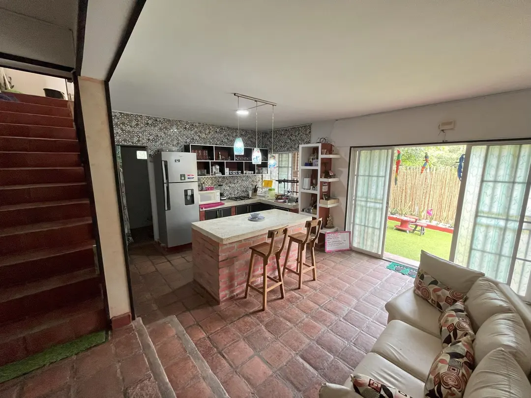 La cocina con isla de ladrillo y taburetes, la escalera al segundo piso y la sala, con puertas de vidrio hacia el jardín.