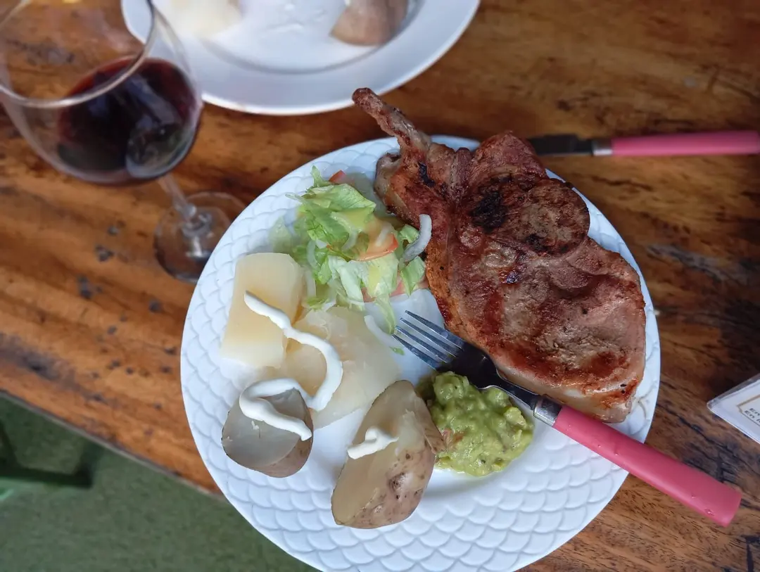 Chuleta a la parrilla con papas, guacamole y ensalada en un plato blanco sobre una mesa de madera.