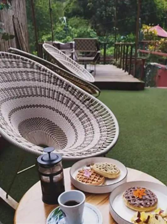 Desayuno en la terraza: sillas tejidas blancas, una prensa francesa de café, waffles y fruta sobre una mesa redonda de madera.