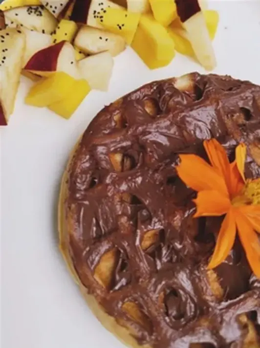 Waffle cubierto de chocolate con trocitos de fruta, decorado con una flor naranja, junto a fruta picada.