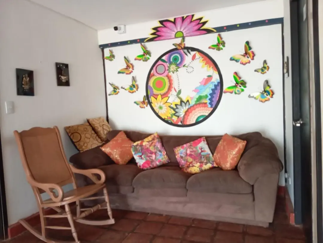 Sala con sofá y cojines de colores, una mecedora de mimbre y un mural de mandala rodeado de mariposas.