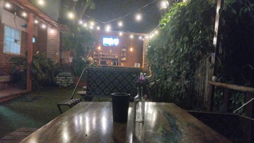 Mesa de madera en el jardín de noche, con un florero; al fondo, guirnaldas de luces, la barra y el tablero de tiza.