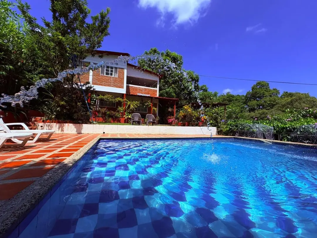 La piscina de Casa Bosco con baldosas azules en damero y un chorro de agua en arco; detrás, la casa de ladrillo de dos pisos bajo un cielo azul.