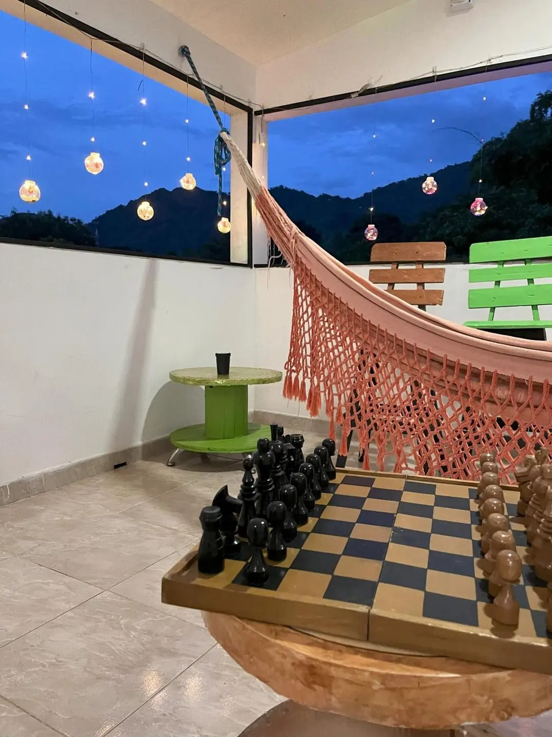 Al anochecer, un tablero de ajedrez grande sobre una base de madera y una hamaca tejida en la terraza, con luces y montañas al fondo.