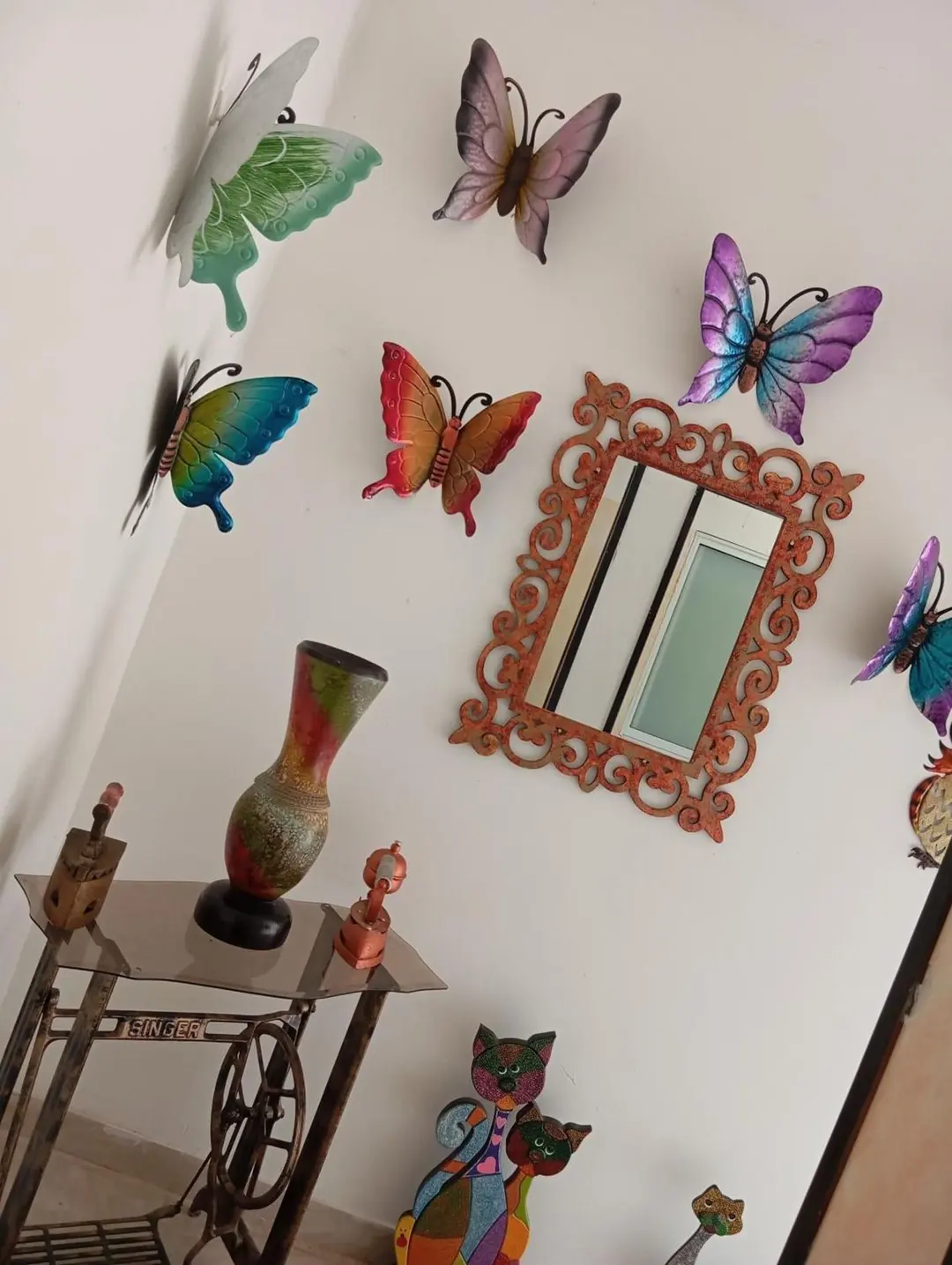 Mariposas de metal de colores alrededor de un espejo con marco tallado en una pared blanca.
